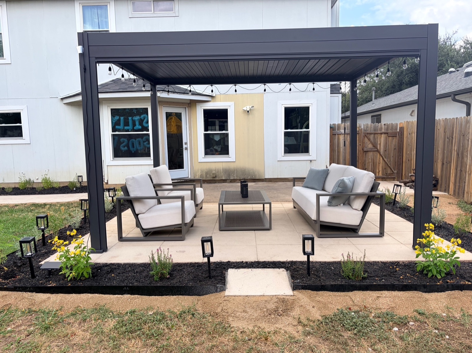 Paver patio extension off the concrete slab with a modern pergola, lounge set, string lights, and flower beds